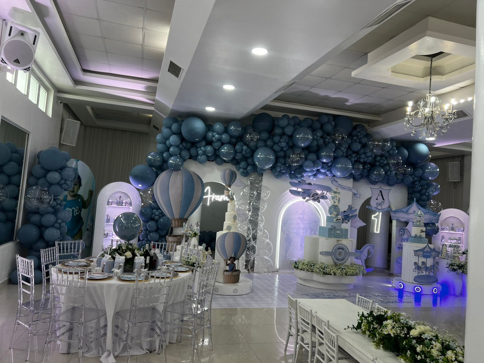 Salón decorado en azul con arco de globos, globos aerostáticos, carrusel, avión y mesas con sillas Tiffany