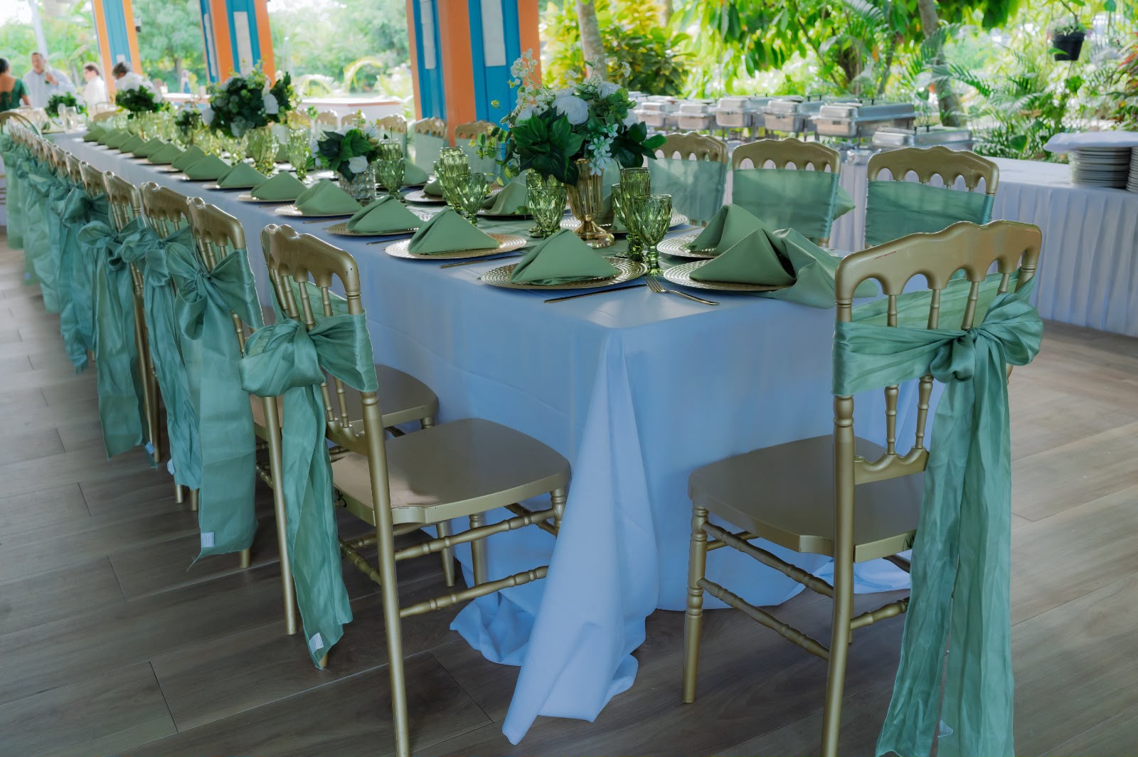 Mesa imperial de boda en Higüey con mantel blanco, servilletas verde salvia, sillas doradas con lazos y flores