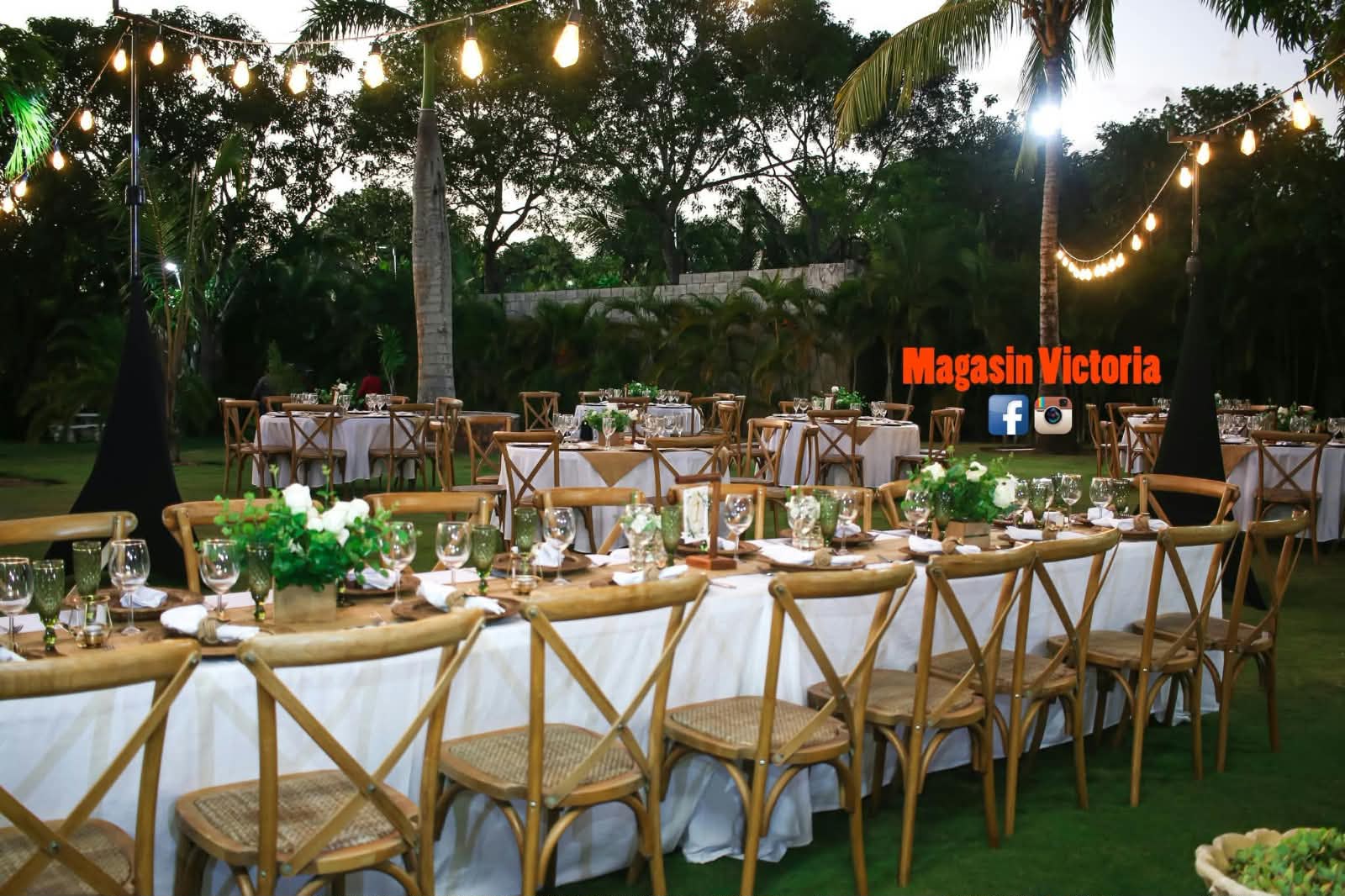 Recepción de boda en jardín con sillas cruzadas de madera, mesas blancas y bombillos colgantes al atardecer