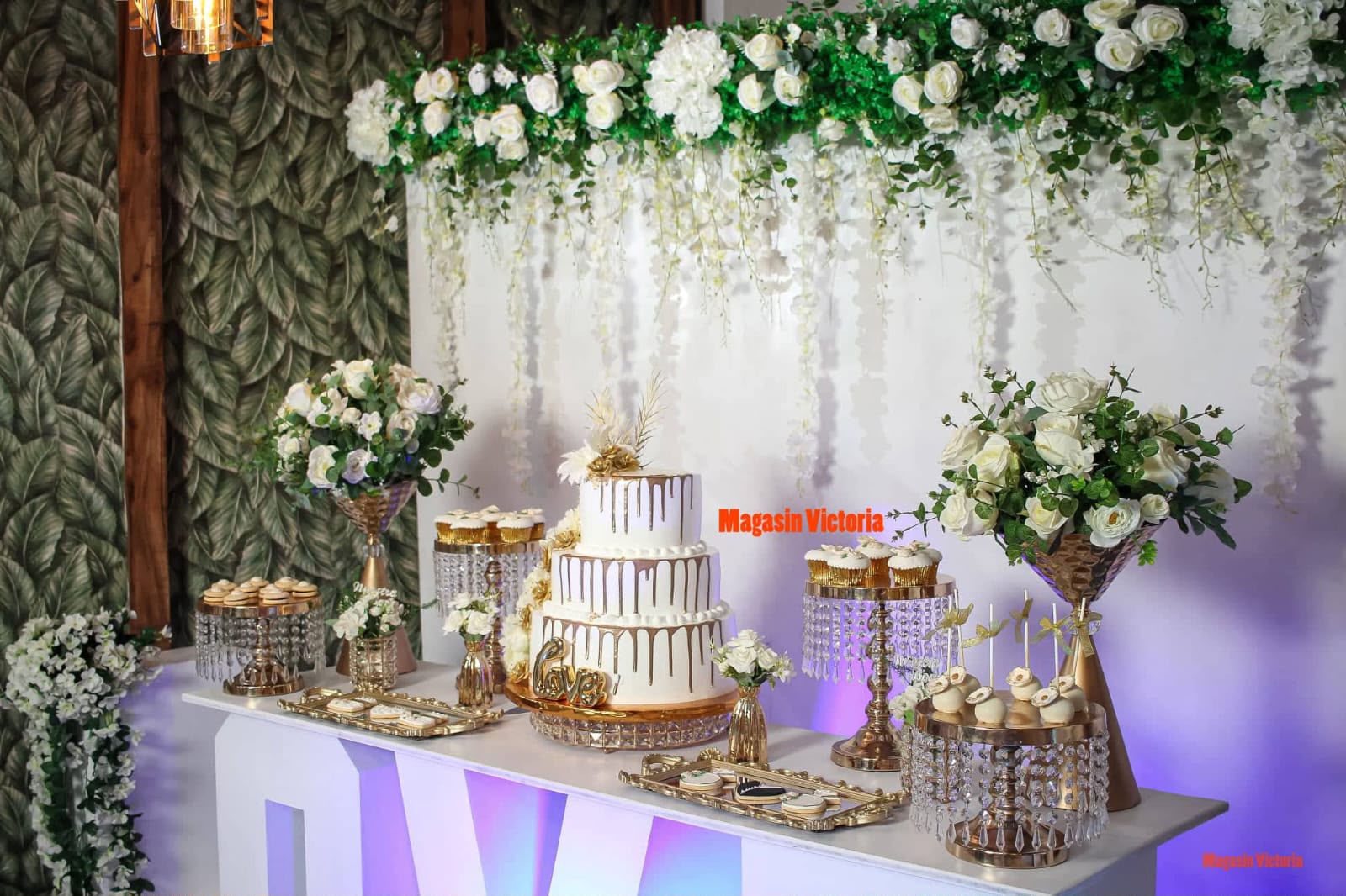Mesa de postres de boda de Magasin Victoria: torta blanca y dorada, letrero Love, cupcakes y cortina de flores blancas