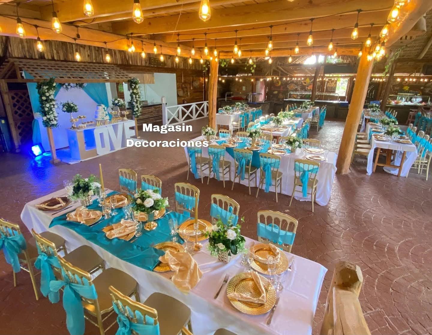 Salón rústico con mesas de boda turquesa y dorado, sillas con lazos, bombillos y letras LOVE al fondo