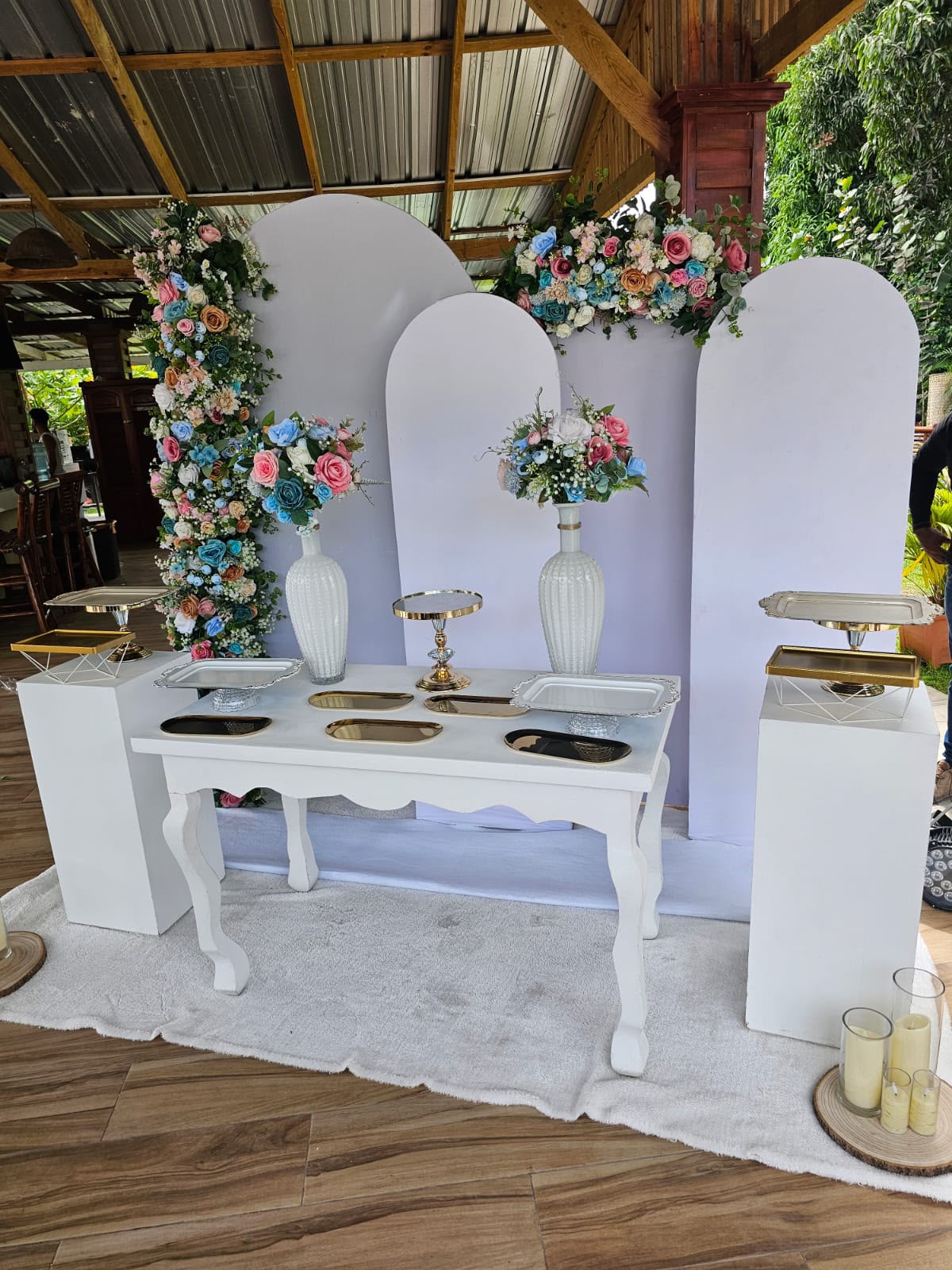 Mesa de postres blanca con paneles arco, columna de flores rosadas y azules, jarrones y bandejas doradas