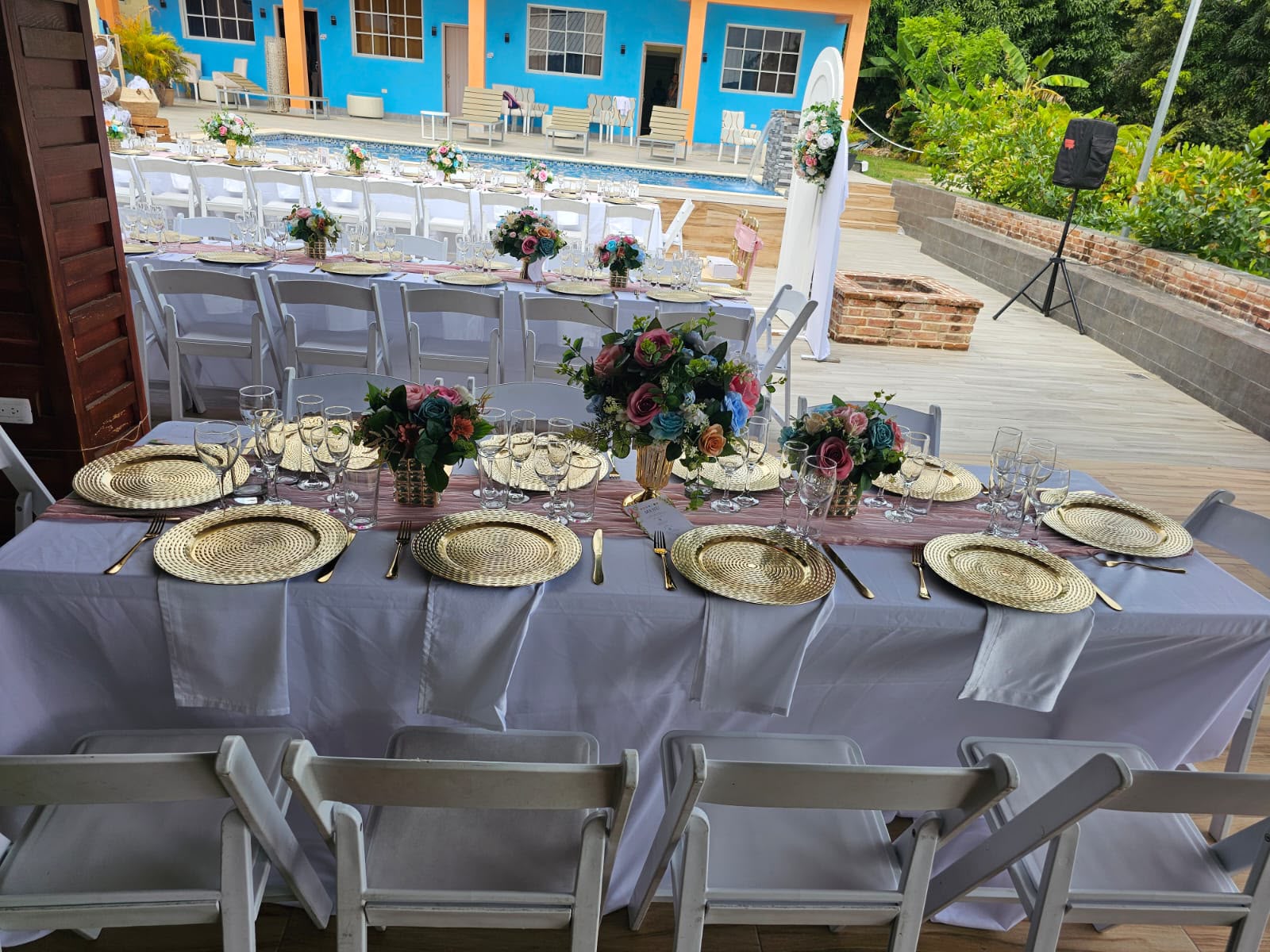 Mesa de boda con bajoplatos dorados, camino rosado y arreglos de rosas; al fondo más mesas junto a la piscina