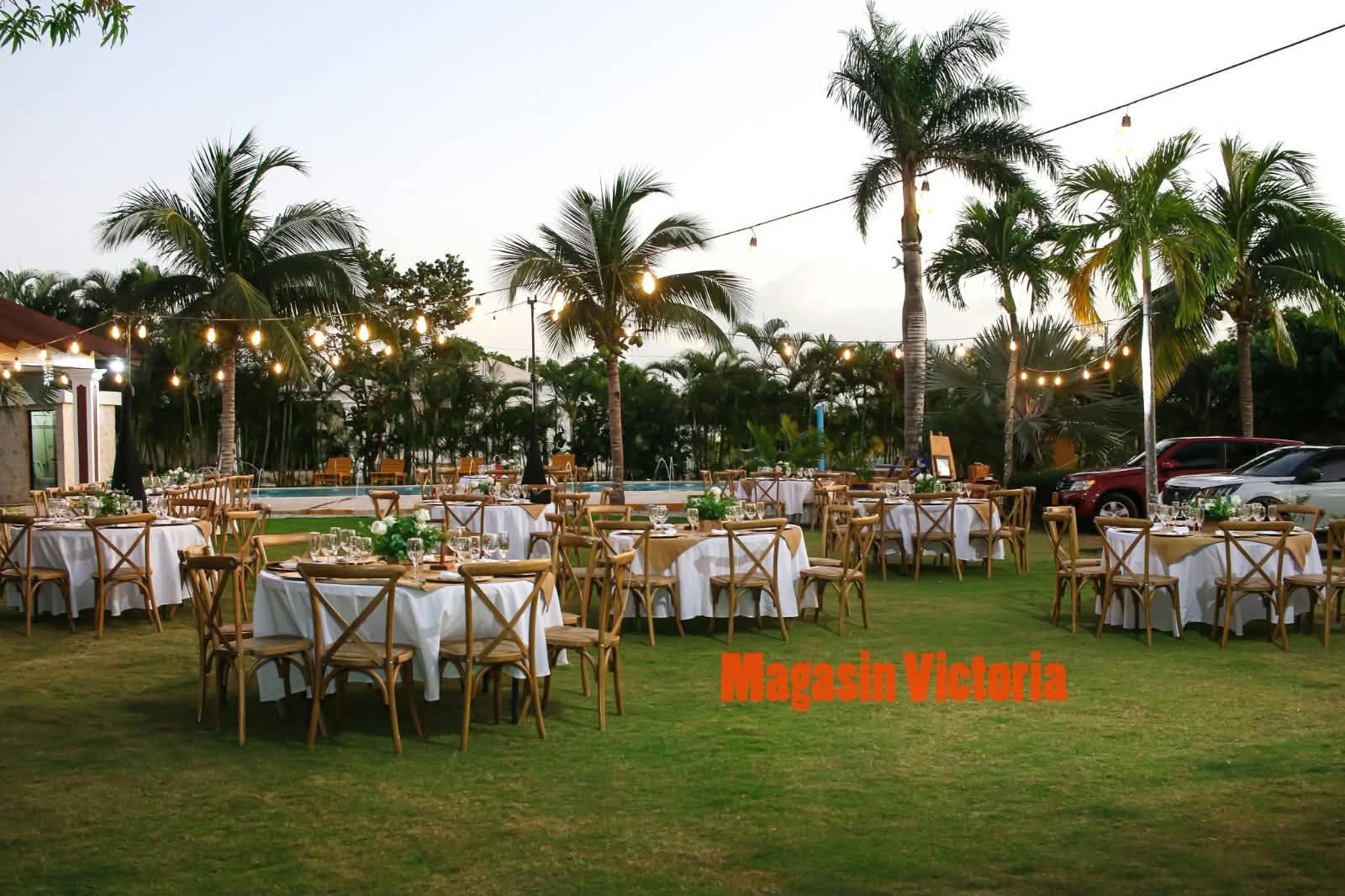 Mesas redondas con sillas crossback de madera, manteles blancos y bombillos entre palmeras, por Magasin Victoria