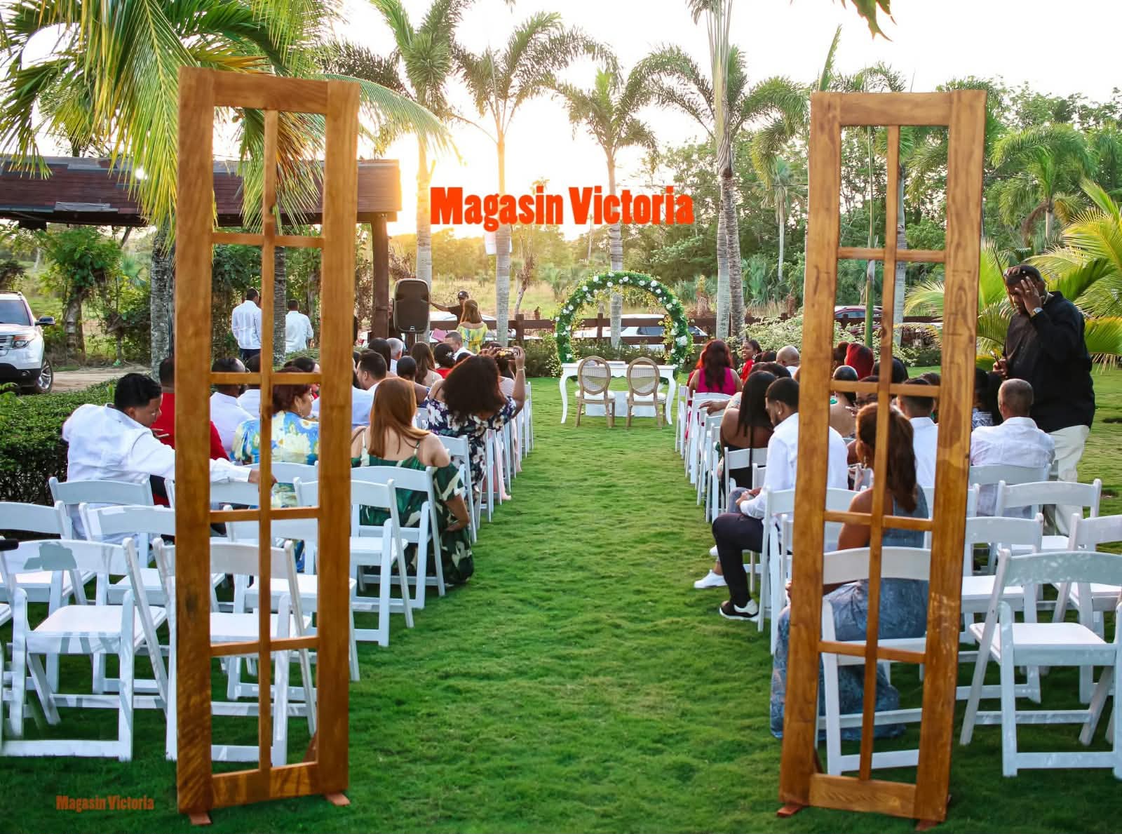Ceremonia de boda al aire libre con puertas de madera, sillas blancas y arco de flores entre palmeras, montada por Magasin Victoria