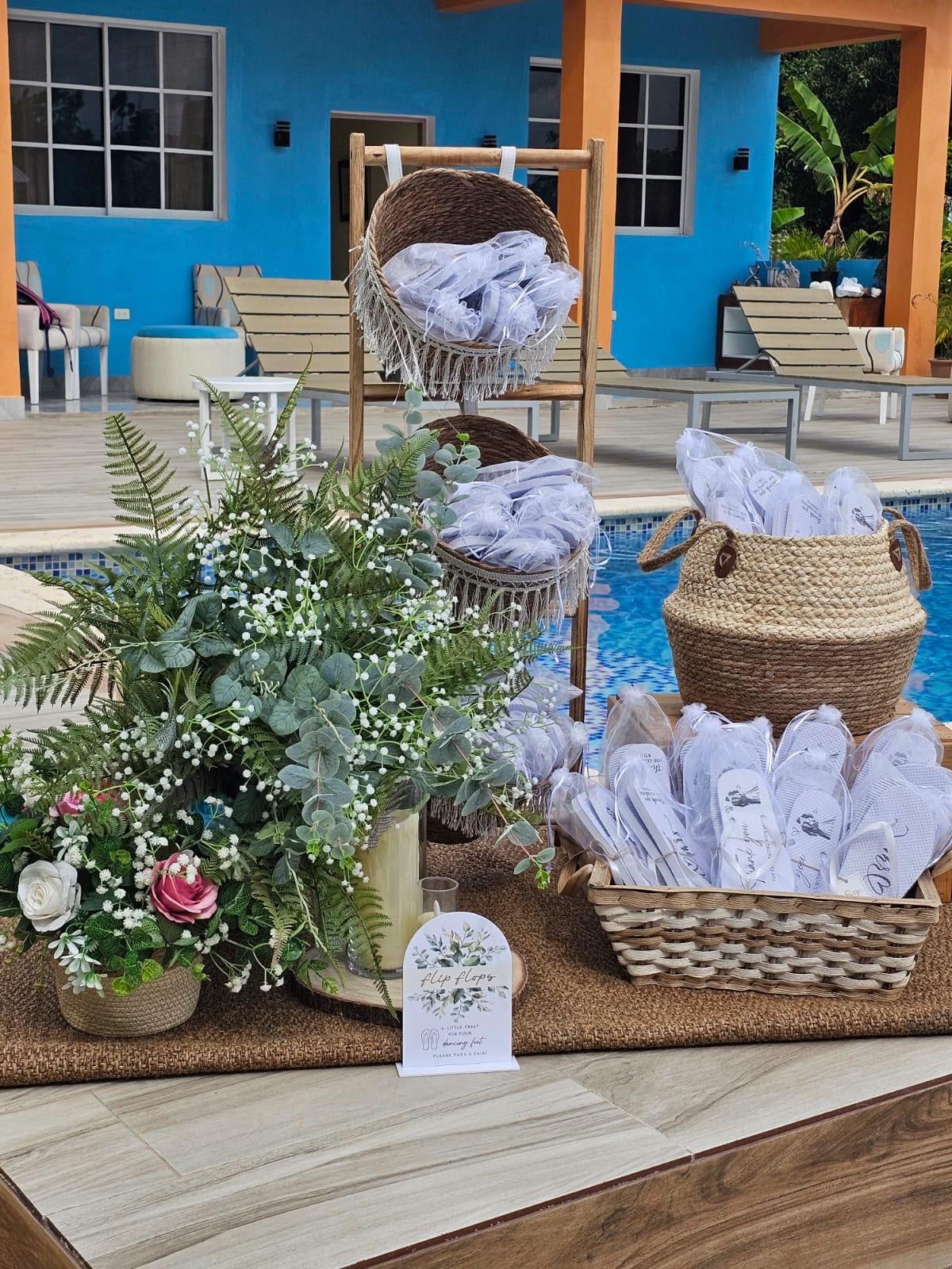 Estación de sandalias para invitados con cestas de mimbre, arreglo de eucalipto y rosas junto a la piscina, por Magasin Victoria