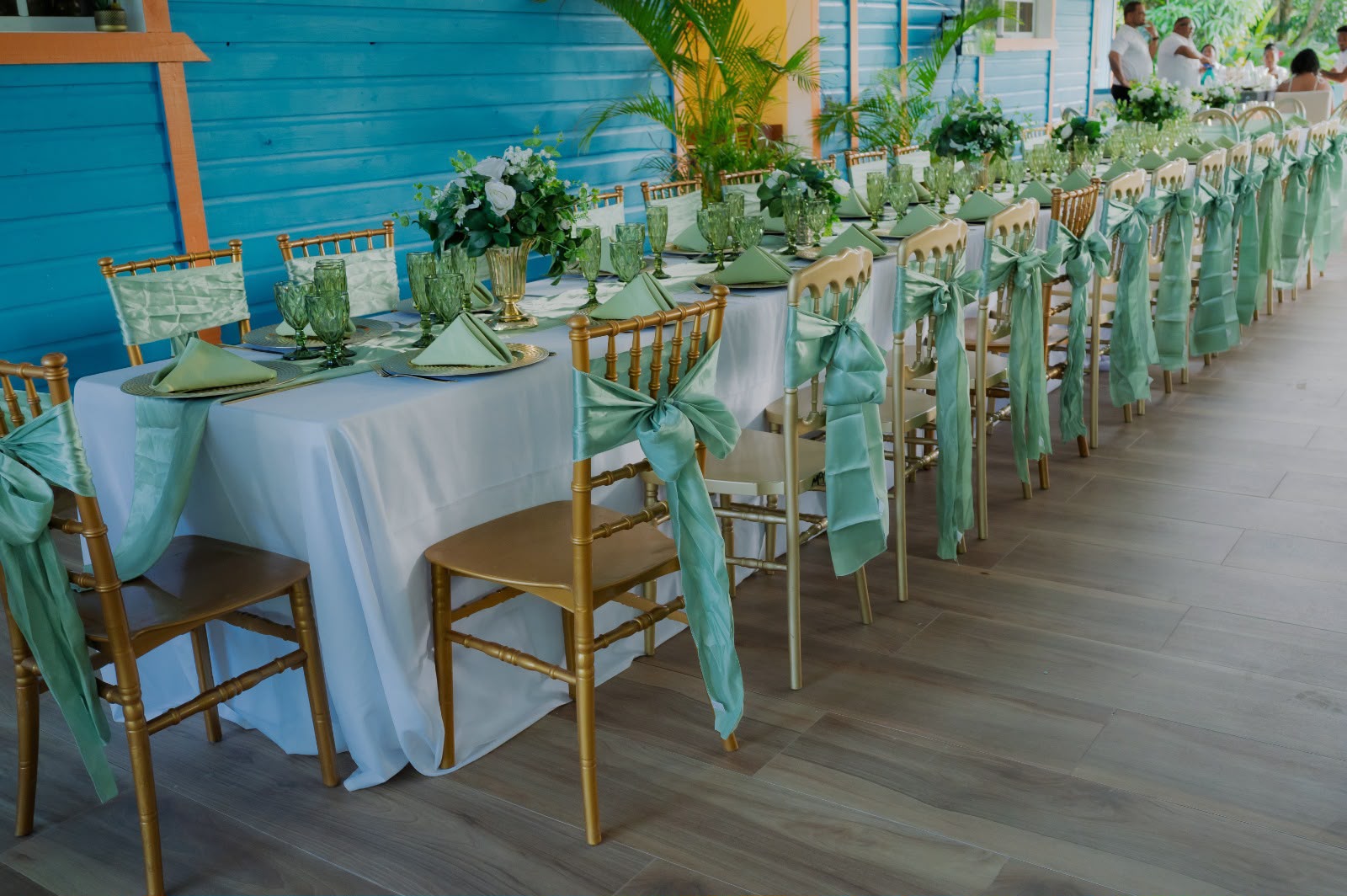 Mesa larga con sillas tiffany doradas y lazos verde menta, copas verdes y centros de flores blancas, montaje de Magasin Victoria
