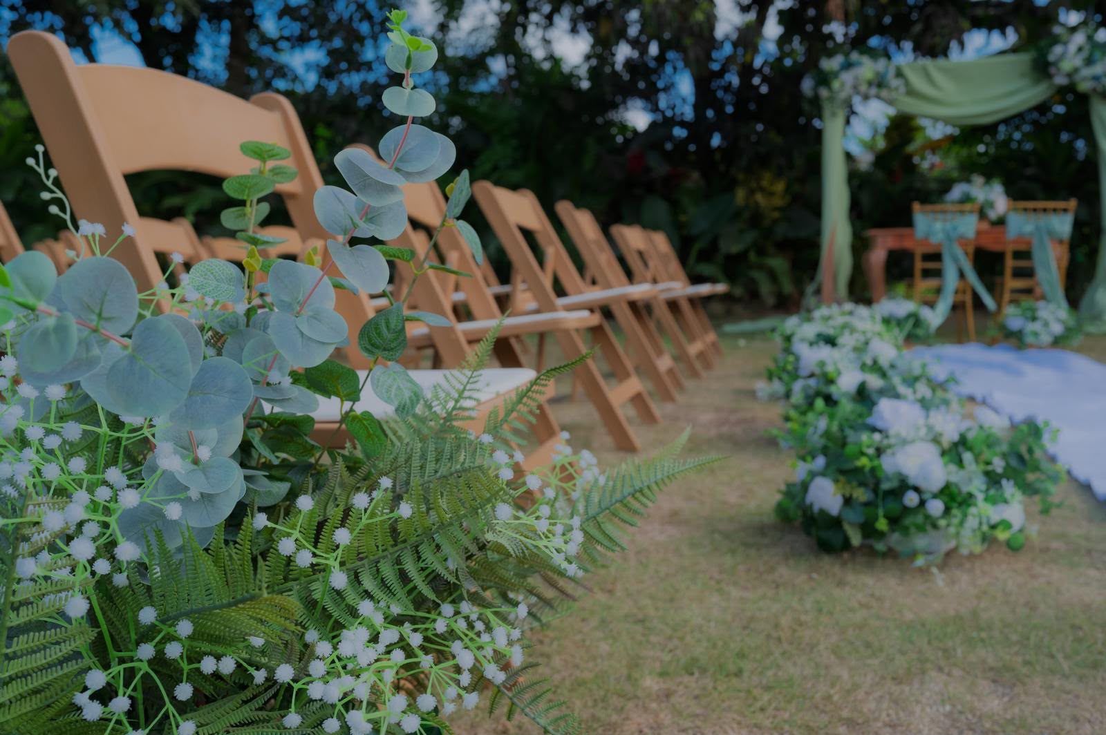Detalle de pasillo de ceremonia con arreglos de eucalipto y flores blancas, sillas de madera y arco verde salvia al fondo