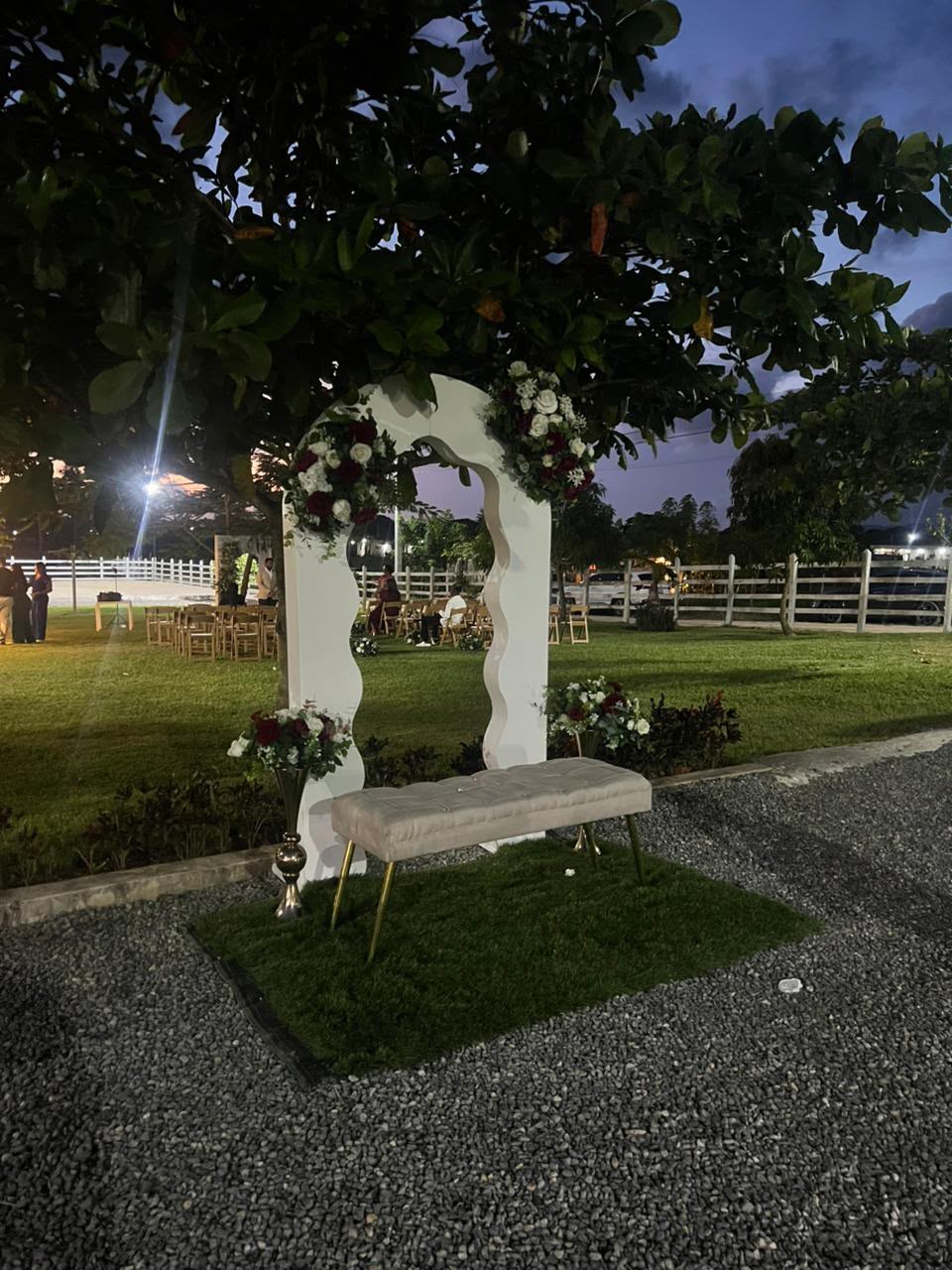 Rincón para fotos con arco blanco ondulado, flores rojas y blancas y banco tapizado en jardín de noche, evento en Higüey