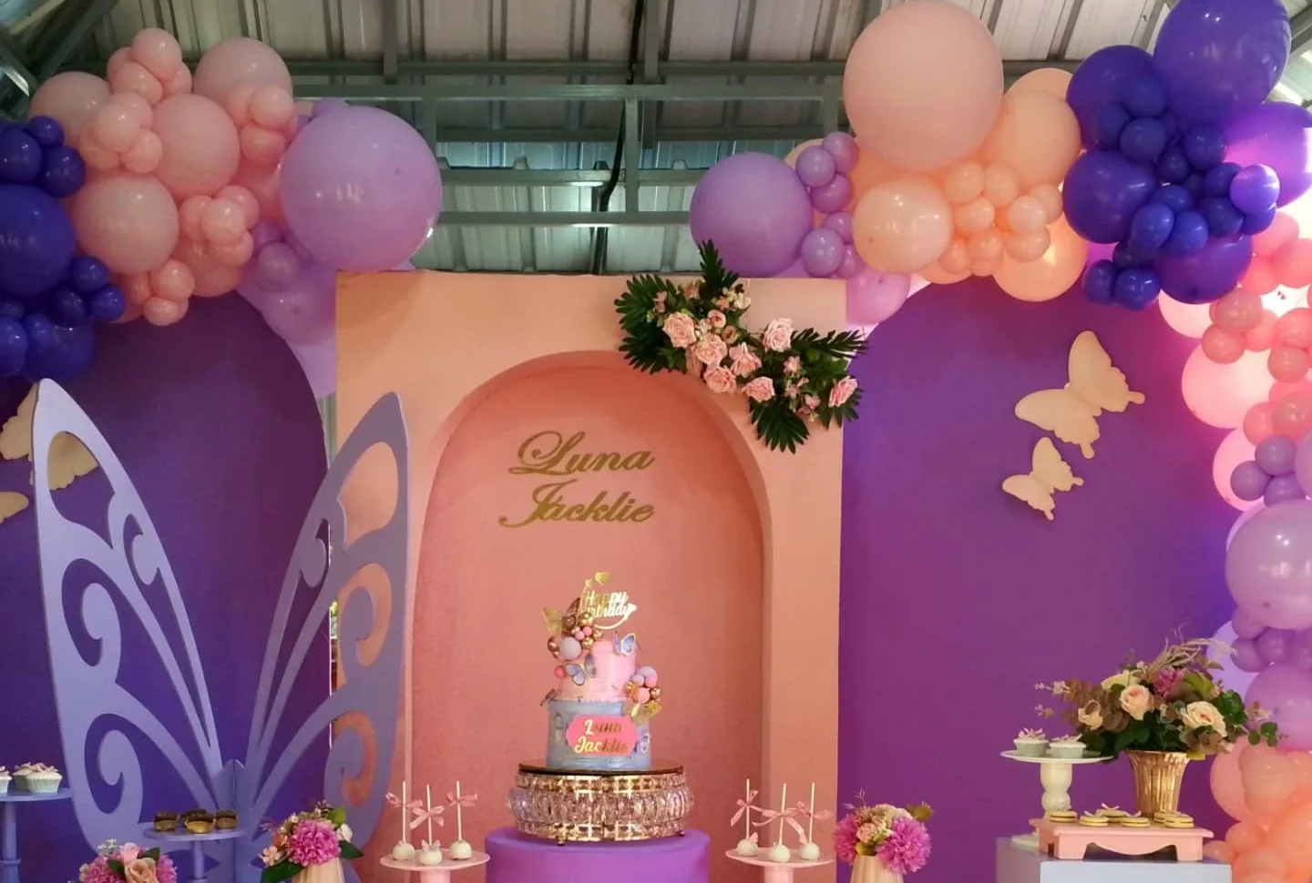 Backdrop de primer cumpleaños con temática de mariposas montado por Magasin Victoria, tonos morado y durazno, arco de globos