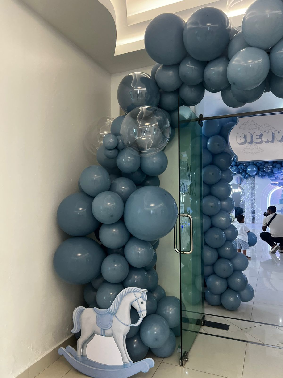 Arco orgánico de globos azul empolvado con burbujas transparentes y caballito de madera en la entrada del salón, por Magasin Victoria
