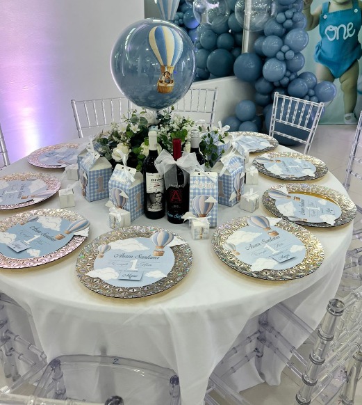 Mesa de primer cumpleaños en tonos azules con globo burbuja de globo aerostático, platos personalizados y pared de globos al fondo, por Magasin Victoria